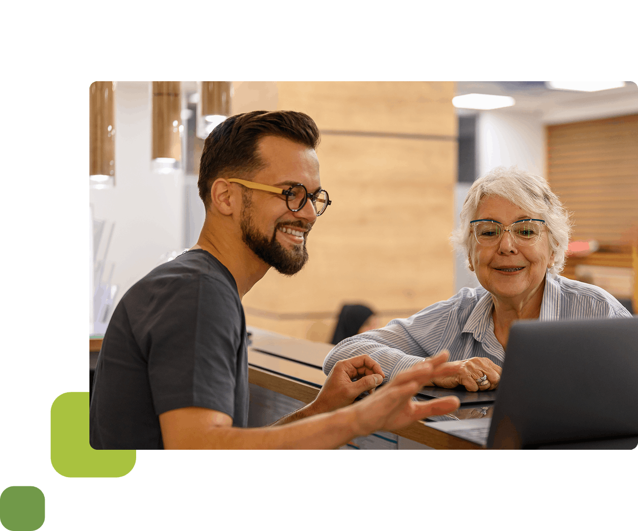 Male receptionist pointing at laptop with elderly woman watching